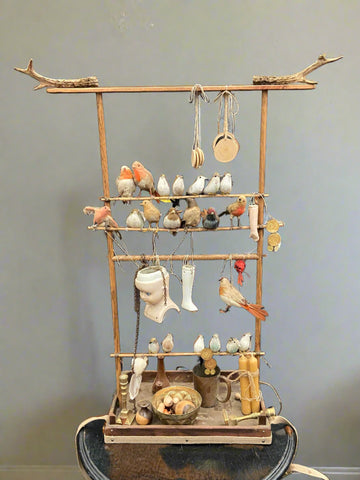 Unusual prop make wooden frame full of odd knick-knacks such as toy birds, candles, a candlestick, animal antlers, and crockery. It looks like it was designed to be worn, for example by a street vendor.