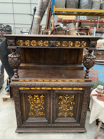 An early English court dresser with a dark vanished wooden base and lighter-coloured inlaid wood. The dresser features many carved details including two large carved acorns which act as supporting pillars and carved foliage details.
