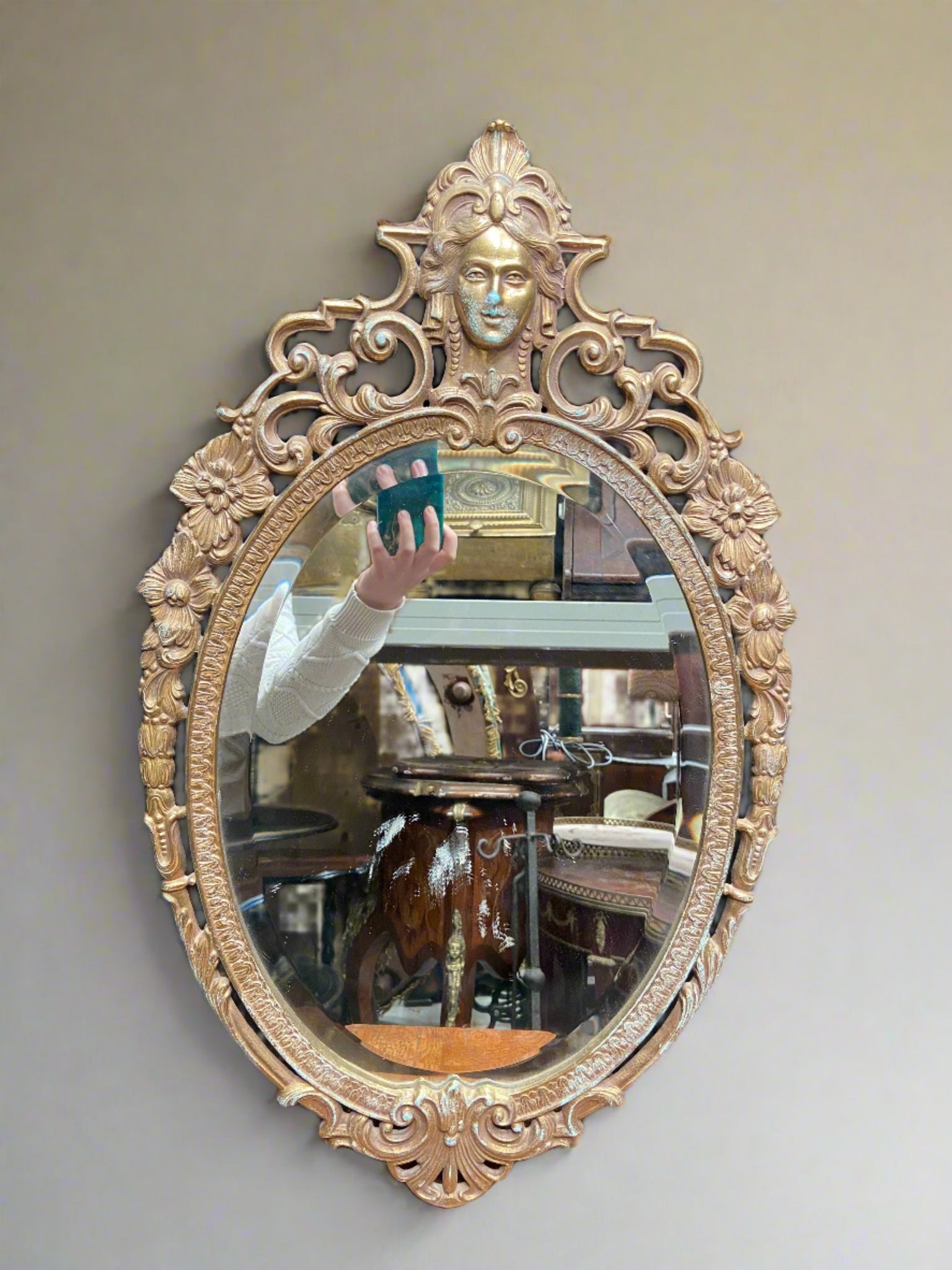 Gold oval Victorian mirror with beveled edging and ornate floral details. Some patina aging on the face.