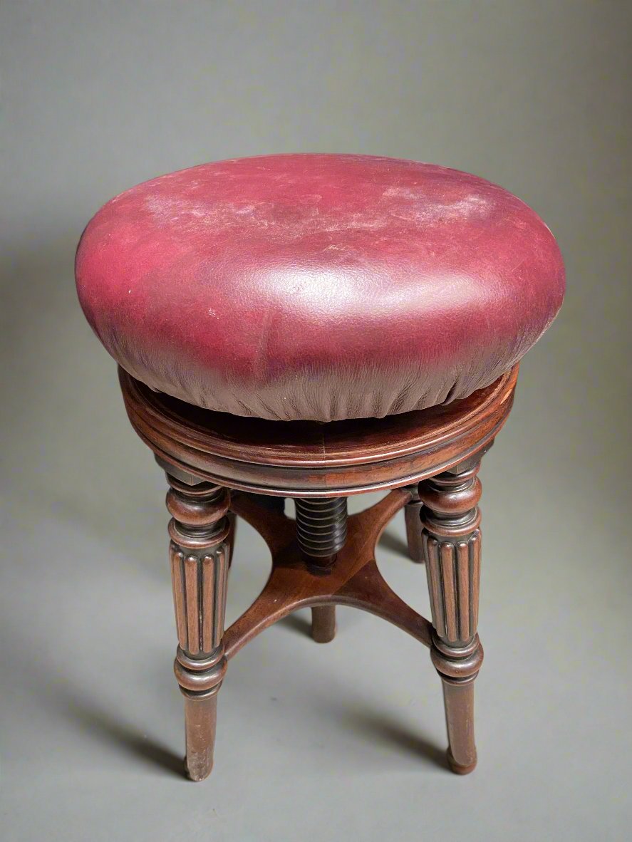 Wooden piano stool upholstered with cherry leather fabric, with an adjustable screw seat. Likely Victorian.