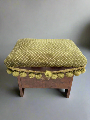 Wooden footstool with olive green and white criss-cross patterned fabric, decorated with pom poms. The seat lifts up to reveal storage.