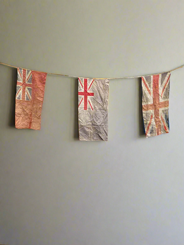 Three large vintage flags, connected by string. Printed onto the fabric are the Union Jack and two Royal Navy Ensign flags.