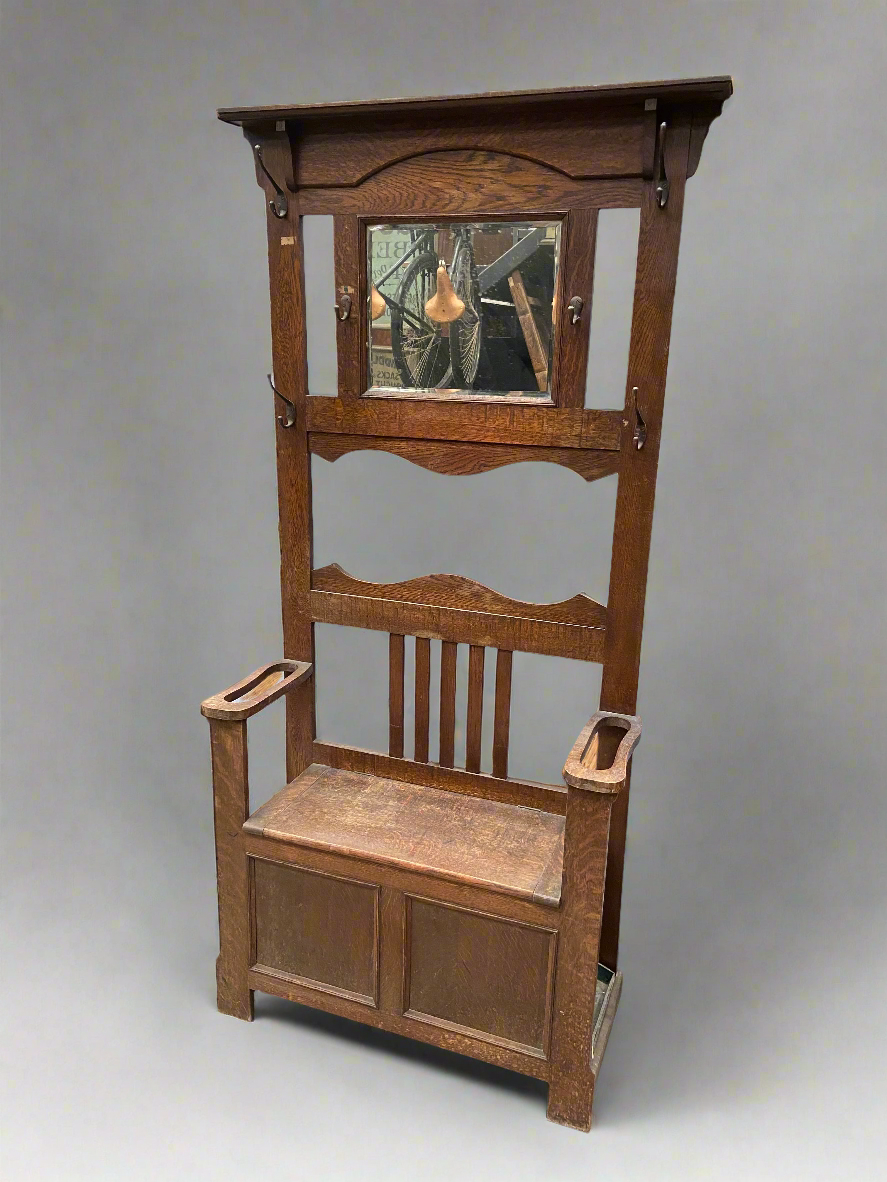 Art Nouveau oak hall stand, circa 1930s. The unit has a bevel edged mirror, six coat hooks, a seat with storage compartment and arm rests with storage for walking sticks.