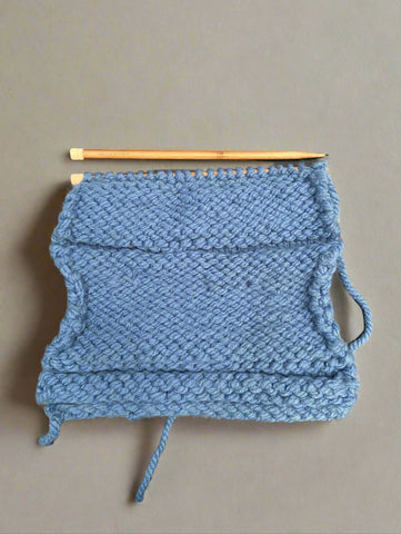 A thick blue wool knitting set, with wooden needles and a square already knitted.