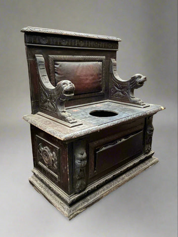 An ornate wooden commode/toilet chair with richly detailed carvings featuring dog heads and a padded leather backrest.