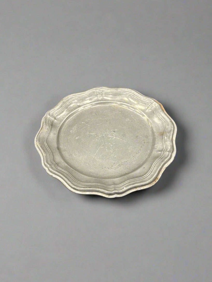 A round, antique, pewter coaster with a ribbed, frilly edge.