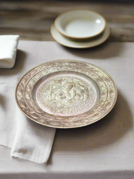 A large antique decorative metal wall plate with latin text and a repoussé design of two figures carrying grapes.