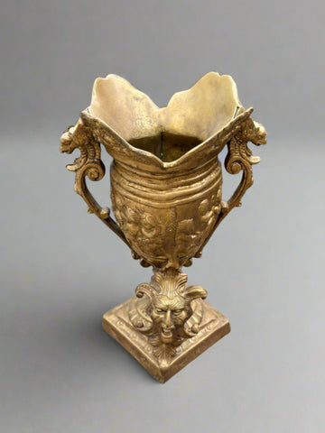 An antique, ornate, brass vase urn with a cast brass joker face at the base and mythical creature handles. Likely French or Italian.