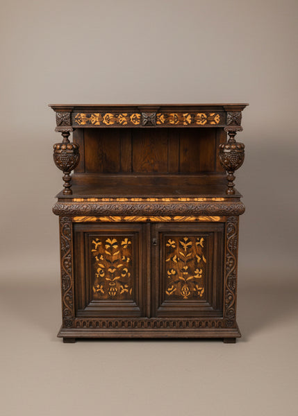 An early English court dresser with a dark vanished wooden base and lighter-coloured inlaid wood. The dresser features many carved details including two large carved acorns which act as supporting pillars and carved foliage details.