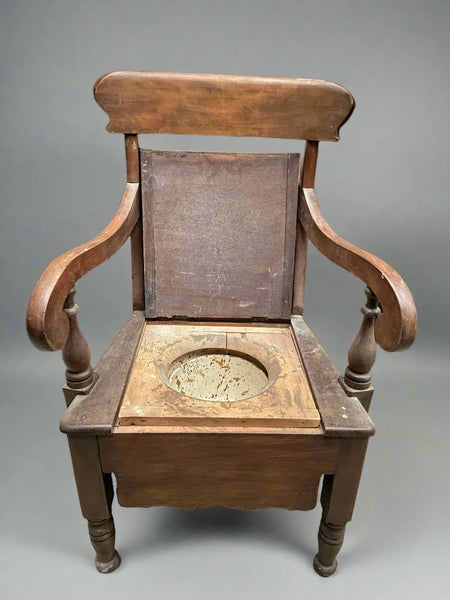 Antique wooden commode with scrolled arms and tall back. The seat panel lifts up to reveal the pot with original wooden lid in place. The inner bowl has had painted effects added to it. Likely Victorian.